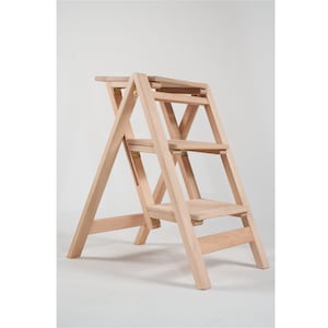 May include: A light-coloured wooden step stool with three steps and a top platform. The step stool folds for storage and has a simple design. The wood has a natural finish, and the hardware is brass-coloured.