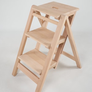 May include: A natural wood step stool with three steps and a top platform. The stool is folded and ready to be used.