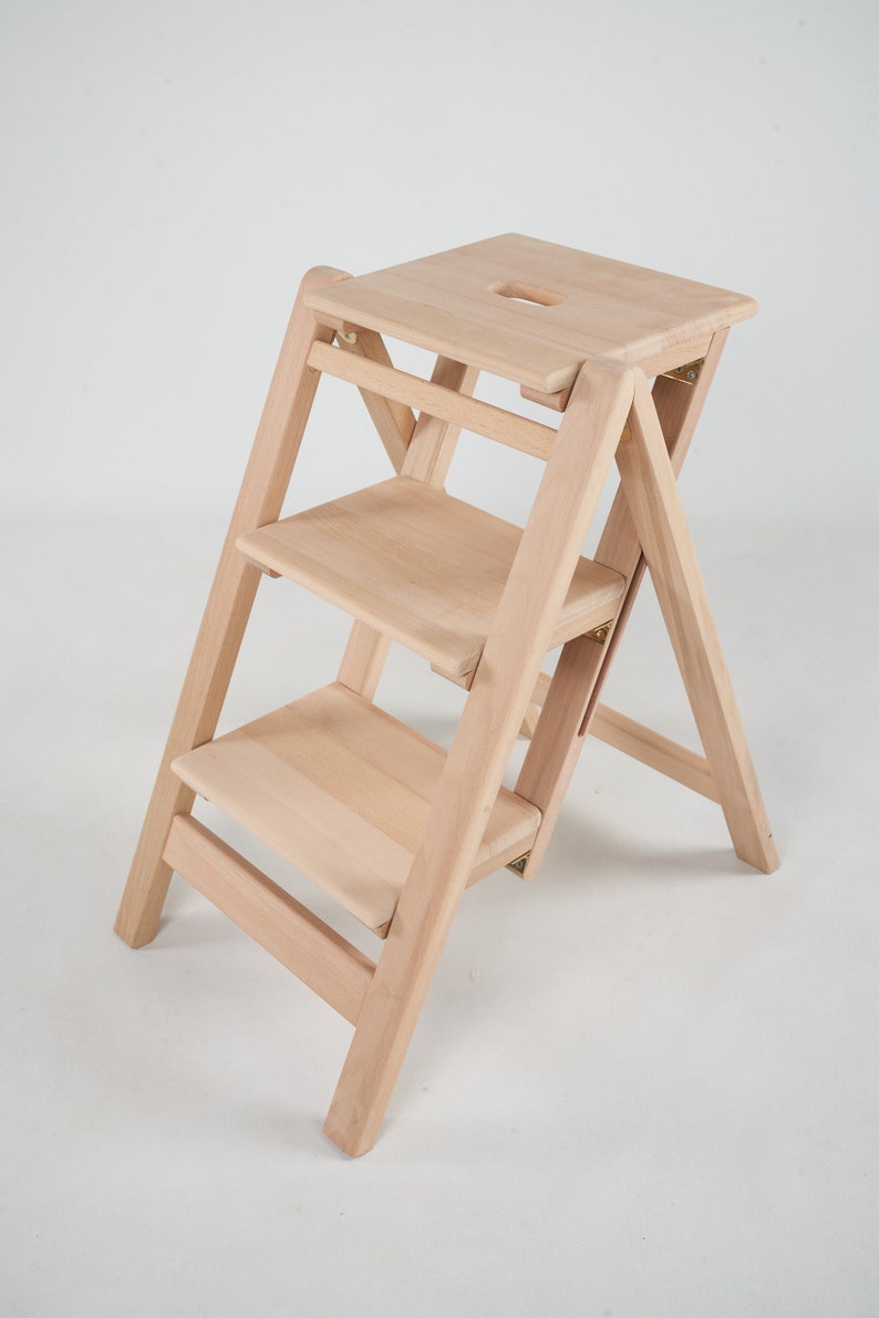 May include: A natural wood step stool with three steps and a top platform. The stool is folded and ready to be used.