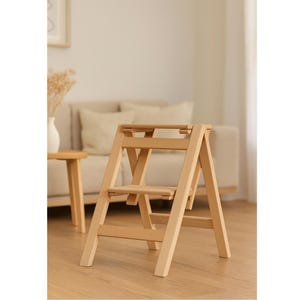 May include: A light-coloured wooden step stool with two steps. The step stool is folded, with the legs angled outwards for stability. The wood has a natural finish, and the design is simple and functional. The height is approximately 60 cm.
