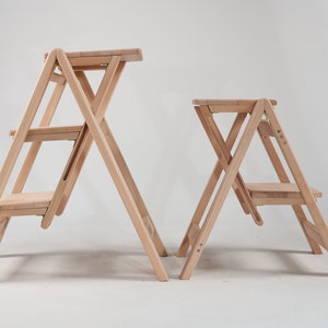 May include: Two wooden step stools, one taller than the other, both with three steps and a foldable design. The stools are made of natural wood and have a light brown finish.