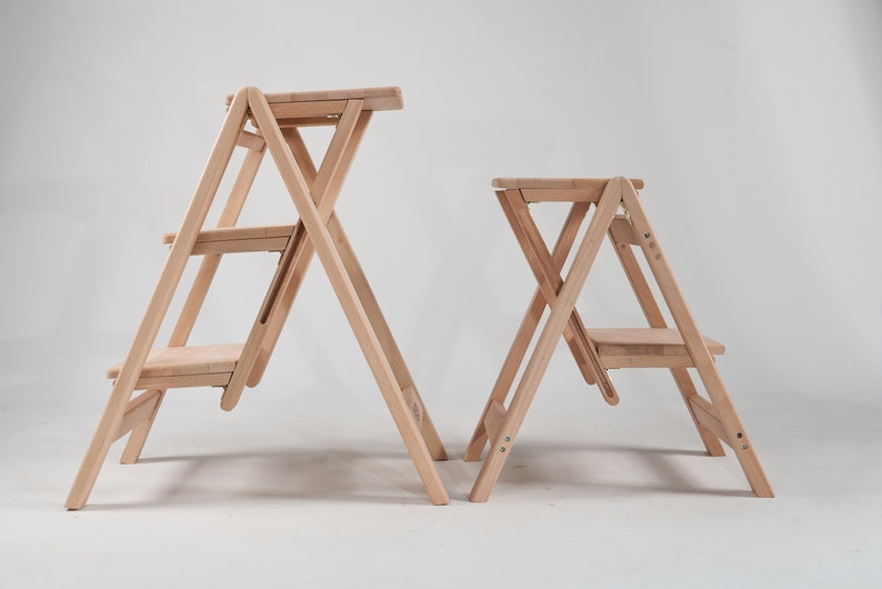 May include: Two wooden step stools, one taller than the other, both with three steps and a foldable design. The stools are made of natural wood and have a light brown finish.