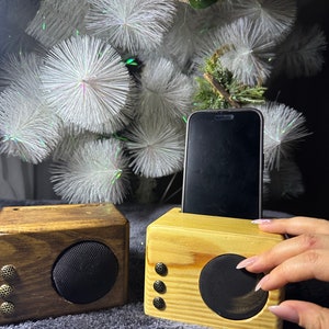 May include: Two wooden Bluetooth speakers, one brown and one natural wood, with a black speaker grille. A smartphone is placed in the top of the natural wood speaker.