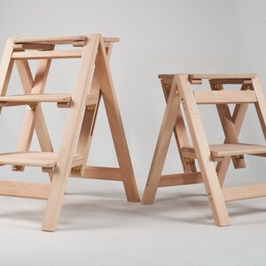 May include: Two wooden step stools, one with three steps and one with two steps, both with a natural finish and gold hinges.