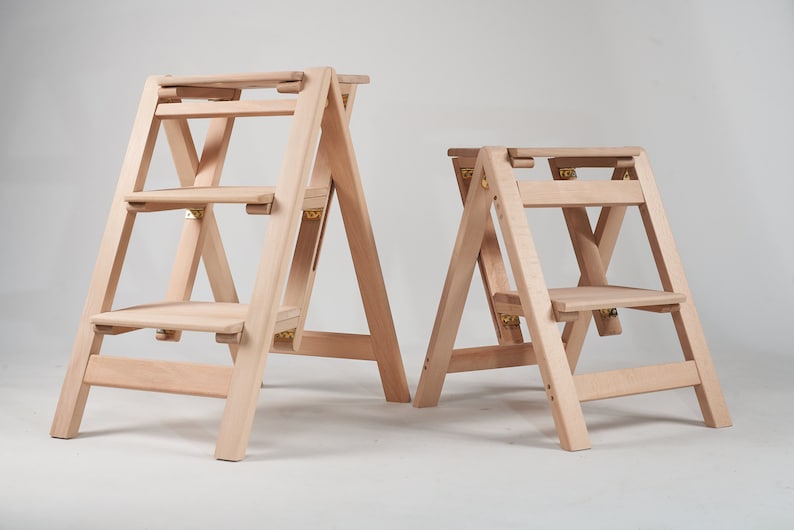 May include: Two wooden step stools, one with three steps and one with two steps, both with a natural finish and gold hinges.