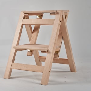 May include: A light wood step stool with two steps and a folding design. The stool is made of natural wood and has a simple, minimalist design.