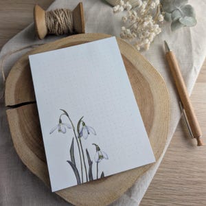 Könnte beinhalten: Ein weißer Notizblock mit einem gepunkteten Muster und einer Aquarell-Illustration von Schneeglöckchen in der rechten unteren Ecke. Der Notizblock liegt auf einer Holzoberfläche.