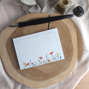 Könnte beinhalten: Ein rechteckiger Notizblock mit floralem Design mit roten Mohnblumen, lila Lavendel und gelben Blumen. Ein schwarzer Kalligraphiestift und eine Tintenflasche liegen auf einer Holzscheibe. Der Notizblock hat einen hellgrünen Rand.