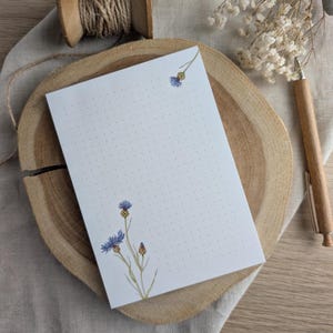 Könnte beinhalten: Ein weißer Notizblock mit blauen und grünen Blumenillustrationen und einem gepunkteten Muster. Der Notizblock liegt auf einer Holzoberfläche.