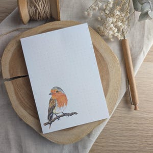 Könnte beinhalten: Ein weißer Notizblock mit einer Amsel-Illustration auf einem Ast. Der Notizblock hat ein gepunktetes Muster und liegt auf einer Holzoberfläche.
