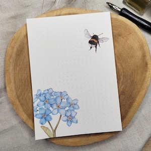 Könnte beinhalten: Ein weißes Papier mit Punktraster und einer Aquarellillustration einer Hummel und blauen Vergissmeinnicht-Blüten. Das Papier liegt auf einem Holztablett mit einem Stift.