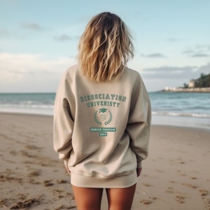 Könnte beinhalten: Ein beiger Pullover mit dem Aufdruck "Dissociation University" und "Honour Program 2024" auf der Rückseite. Der Pullover wird von einer Person getragen, die an einem Strand steht.