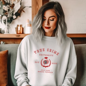 Peut inclure: Un sweat-shirt gris clair avec le texte "True Crime University" en rouge. Le sweat-shirt présente un graphique rouge d'un bonnet de graduation, d'un diplôme et d'une couronne de laurier avec le texte "Est 2024" et "Cold Case Honors Program".