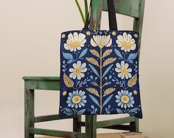 May include: A blue canvas tote bag with a floral pattern in white, yellow, and gold. The bag is hanging on a green wooden chair. There are pears and a pitcher on the floor in front of the chair.