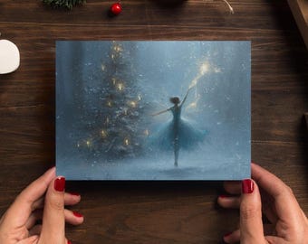 May include: A greeting card featuring a ballerina in a blue dress, arms outstretched, in a snowy scene with a Christmas tree. The artwork has a soft, dreamy quality with a focus on the dancer and the festive background.