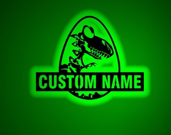 May include: A green glow-in-the-dark dinosaur skeleton inside an egg with the text "CUSTOM NAME" below. The image is set in a dark room with a desk, a computer, and a lamp.