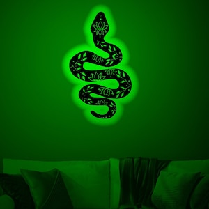 Könnte beinhalten: Eine schwarze Metall-Schlangen-Wanddekoration mit floralem Muster, die in grünem Licht vor einer grünen Wand beleuchtet wird. Die Schlange hängt über einem Sofa mit Kissen.