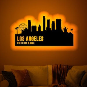 May include: A black silhouette of the Los Angeles skyline with the text "LOS ANGELES" and "CUSTOM NAME" below. The silhouette is illuminated with a warm orange glow and is displayed on a wall in a living room setting.
