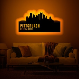 Puede incluir: Una silueta negra de metal del horizonte de Pittsburgh con el texto "PITTSBURGH" y "CUSTOM NAME" debajo. La silueta está iluminada con un cálido resplandor naranja y está montada en una pared en un ambiente de sala de estar.