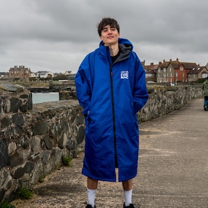 Puede incluir: Una persona lleva una toalla con capucha azul con el texto "Coast Mac" en la parte delantera. La toalla está cerrada con cremallera y la persona está de pie en un camino de piedra.