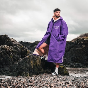Puede incluir: Una persona con un abrigo largo con capucha morado con un logotipo blanco, de pie sobre rocas cerca del océano. El abrigo está abierto, revelando un forro color crema y pantalones cortos negros. La persona lleva calcetines blancos y sandalias.