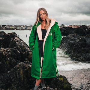 Puede incluir: Un abrigo largo de color verde brillante con capucha e interior color crema. El abrigo lo lleva una persona de pie en una playa rocosa. El abrigo tiene un logotipo en el pecho y está abrochado con cremallera. La persona lleva zapatos negros.