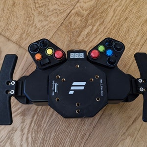 Könnte beinhalten: Schwarzes Rennlenkrad mit einem digitalen Display, das 8,88 anzeigt, einem weißen F-Logo und zwei schwarzen Schaltwippen. Das Lenkrad hat mehrere Knöpfe und ein Farbkonzept in Rot, Gelb, Grün und Blau.