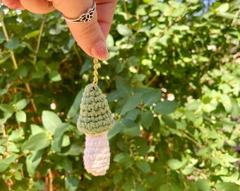 May include: A handmade crochet mushroom charm with a green cap and white stem, suspended by a gold chain. The mushroom is held against a backdrop of green foliage, showcasing its detailed texture and color contrast.