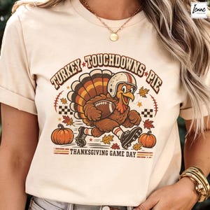 Pode incluir: Uma t-shirt cor de creme com um gráfico de um peru a usar um capacete de futebol americano e chuteiras enquanto segura uma bola. O texto diz TURKEY TOUCHDOWNS PIE e THANKSGIVING GAME DAY, rodeado por abóboras cor de laranja e folhas de outono.