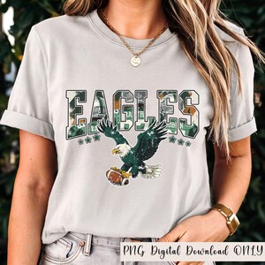 May include: A light gray t-shirt featuring the word "EAGLES" in a camouflage design with green accents. Below the text is an illustration of an eagle holding a football. The text "PNG Digital Download ONLY" is at the bottom.