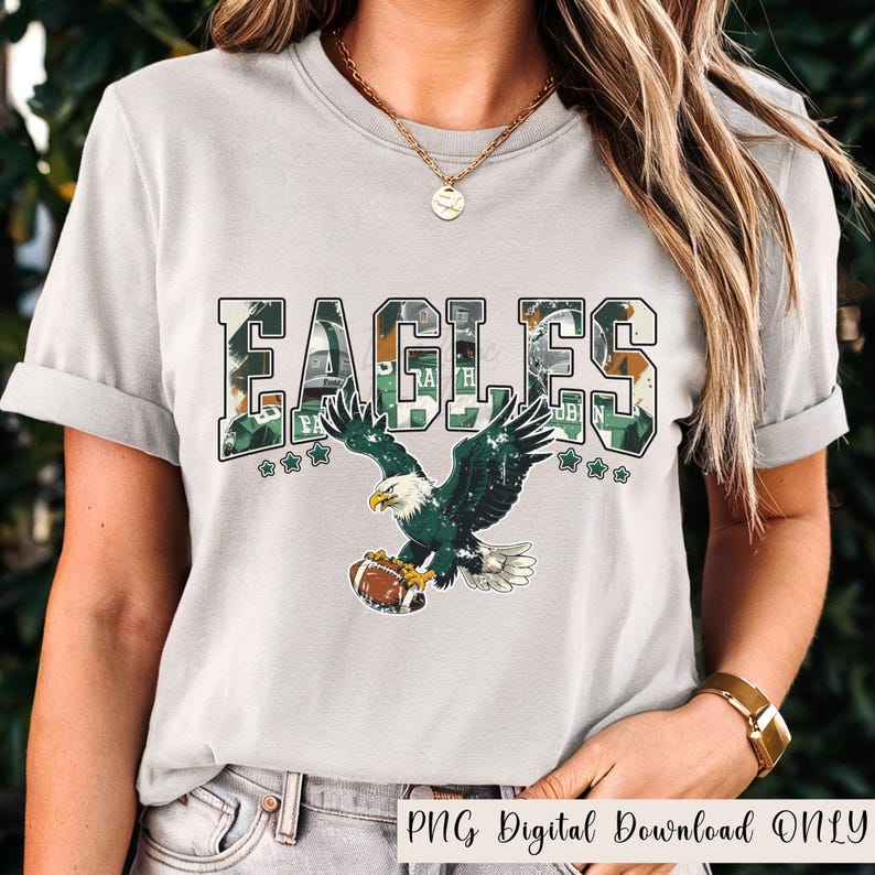May include: A light gray t-shirt featuring the word "EAGLES" in a camouflage design with green accents. Below the text is an illustration of an eagle holding a football. The text "PNG Digital Download ONLY" is at the bottom.