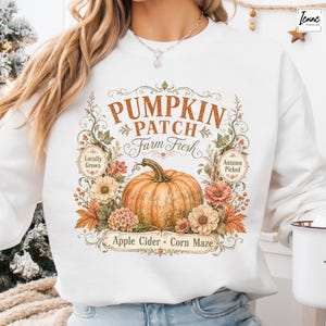 May include: A white crewneck sweatshirt featuring a graphic print of a large orange pumpkin surrounded by autumn flowers and leaves. The text reads PUMPKIN PATCH, Farm Fresh, Locally Grown, Autumn Picked, Apple Cider, and Corn Maze in various fonts.
