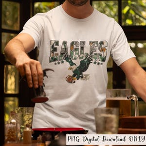 May include: White t-shirt featuring the word "EAGLES" in a camouflage design, with a green and white eagle graphic holding a football. The shirt has the text "PNG Digital Download ONLY" at the bottom.