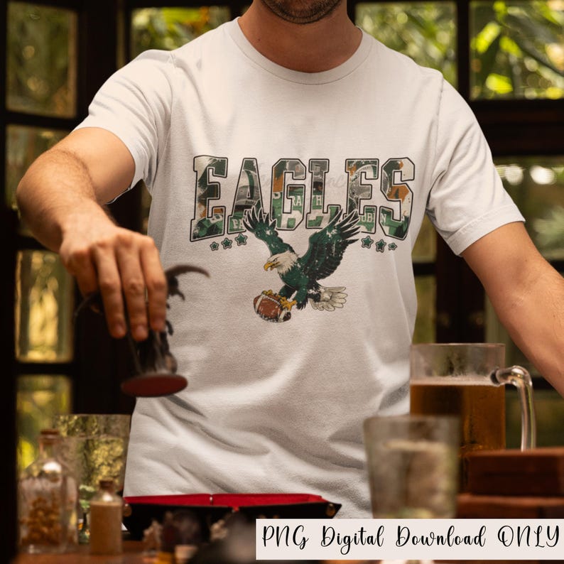 May include: White t-shirt featuring the word "EAGLES" in a camouflage design, with a green and white eagle graphic holding a football. The shirt has the text "PNG Digital Download ONLY" at the bottom.