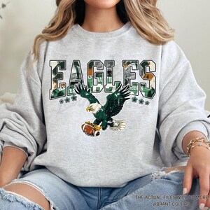 May include: A light gray sweatshirt featuring the word "EAGLES" in a green and white camouflage design. Below the word is a graphic of a green and white eagle clutching a football. The text "THE ACTUAL FILES WILL BE MORE VIBRANT COLORS" is at the bottom.