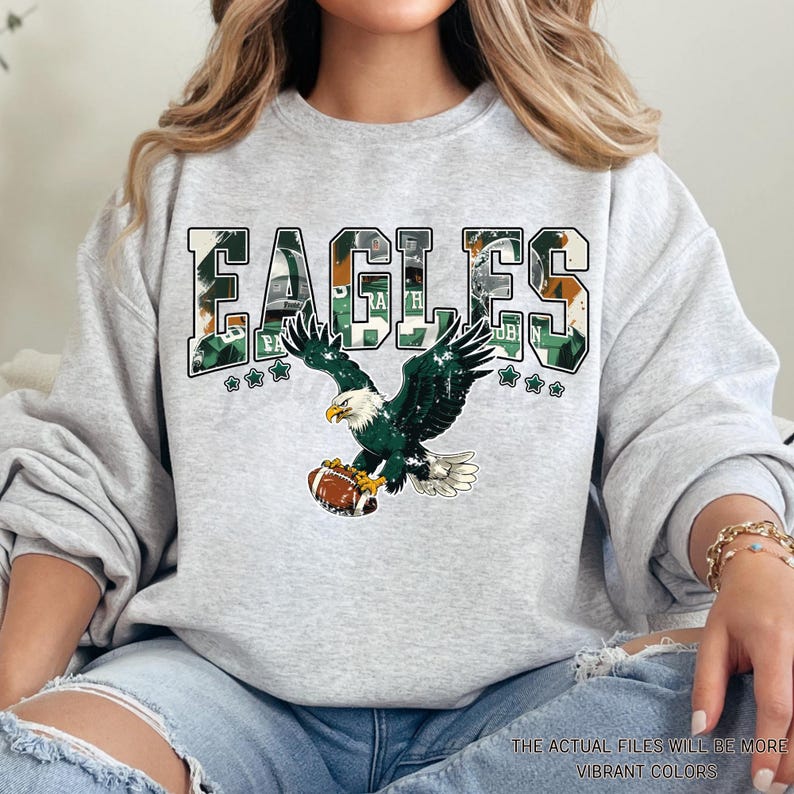 May include: A light gray sweatshirt featuring the word "EAGLES" in a green and white camouflage design. Below the word is a graphic of a green and white eagle clutching a football. The text "THE ACTUAL FILES WILL BE MORE VIBRANT COLORS" is at the bottom.