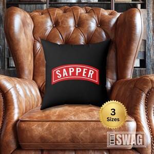 May include: Black throw pillow with a red and white "SAPPER" patch. The pillow is on a brown leather armchair.