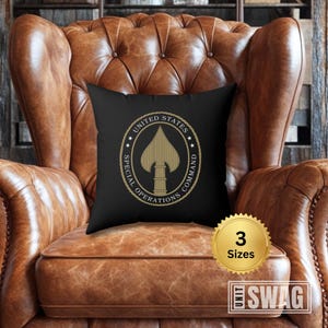 May include: Black throw pillow with a gold United States Special Operations Command logo. The logo features a spearhead with a star on each side. The pillow is sitting on a brown leather armchair.
