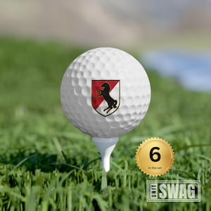 May include: A white golf ball with a black and red crest featuring a rearing horse. The crest is surrounded by a gold border. The golf ball is on a tee and there is a gold sticker with the number 6 and the text "in the set".
