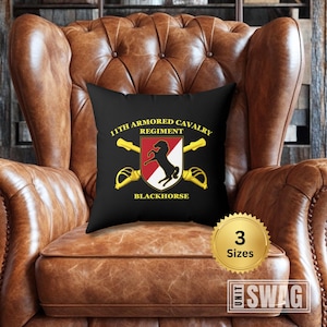 May include: Black throw pillow with white text that reads "11th Armored Cavalry Regiment Blackhorse" and a red and white shield with a black horse. The pillow is on a brown leather armchair.
