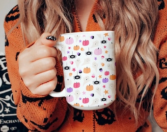 Op de afbeelding: Een witte keramische mok met een Halloween-thema. De mok heeft een herhalend patroon van pompoenen, geesten, spinnen, snoepmais en sterren in roze, oranje, paars en zwart.