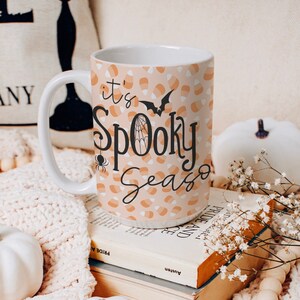 Puede incluir: Una taza de cerámica blanca con un patrón de maíz dulce de color melocotón y blanco. La taza tiene un asa negra y dice "it's Spooky Season" en texto negro con un gráfico de araña y murciélago.