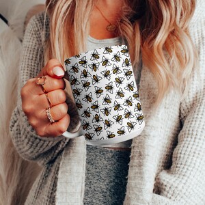 May include: White ceramic mug with a black and yellow bee pattern. The mug is held by a person's hand.