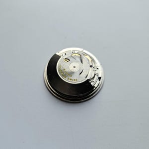 K&ouml;nnte beinhalten: Nahaufnahme eines Uhrwerks mit komplexen Zahnr&auml;dern und Komponenten. Das kreisf&ouml;rmige Uhrwerk hat eine schwarz-silberne Farbgebung. Der Text "17 Jewels Swiss" ist auf dem Uhrwerk zu sehen.