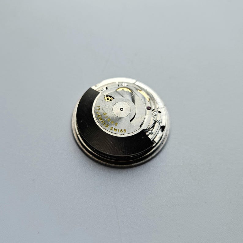 K&ouml;nnte beinhalten: Nahaufnahme eines Uhrwerks mit komplexen Zahnr&auml;dern und Komponenten. Das kreisf&ouml;rmige Uhrwerk hat eine schwarz-silberne Farbgebung. Der Text "17 Jewels Swiss" ist auf dem Uhrwerk zu sehen.