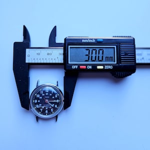May include: A black digital caliper measuring a vintage Timex watch face. The watch face is black with white numerals and hands. The caliper reads 30.0 mm, which is approximately 1.18 inches. The watch face is held between the caliper jaws.