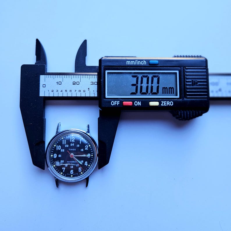 May include: A black digital caliper measuring a vintage Timex watch face. The watch face is black with white numerals and hands. The caliper reads 30.0 mm, which is approximately 1.18 inches. The watch face is held between the caliper jaws.