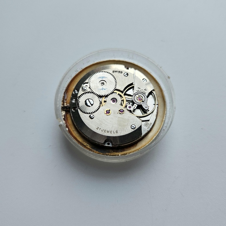 Może przedstawiać: Zbliżenie mechanizmu zegarka z widocznymi kołami zębatymi, kamieniami i tekstem "SWISS" i "21 JEWELS". Mechanizm jest zamknięty w przezroczystej, okrągłej plastikowej obudowie. Metalowe elementy mają srebrne wykończenie.