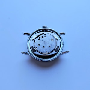 May include: A disassembled wristwatch, showcasing the internal mechanism. The watch's silver casing surrounds the intricate, circular movement, revealing gears, screws, and other components. The watch's lugs extend outward, ready for a strap. The background is a light blue.