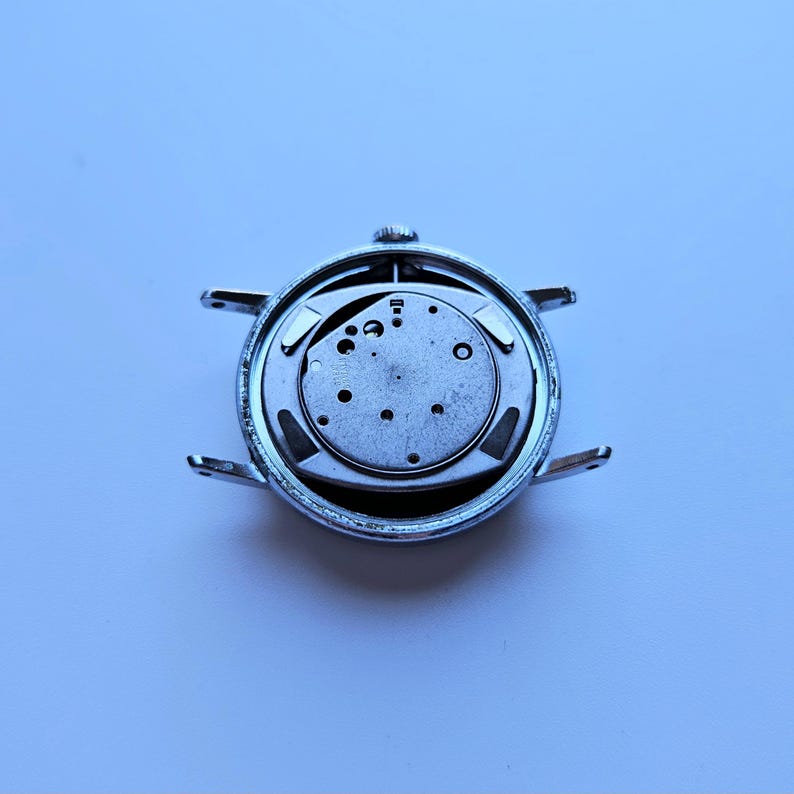May include: A disassembled wristwatch, showcasing the internal mechanism. The watch's silver casing surrounds the intricate, circular movement, revealing gears, screws, and other components. The watch's lugs extend outward, ready for a strap. The background is a light blue.